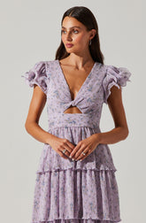 A woman is wearing a light purple dress with a floral pattern. The dress features puffed, ruffled short sleeves, a deep V neckline with a twist knot at the bust, and a cutout detail just below the knot. The bodice and skirt have a layered, tiered design with ruffles and vertical pleats, creating a textured, flowing appearance.